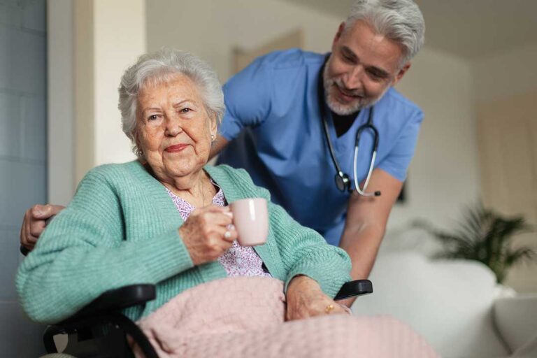 Two older adults and a caregiver discuss daily needs while seated at a dining table with paperwork and coffee.