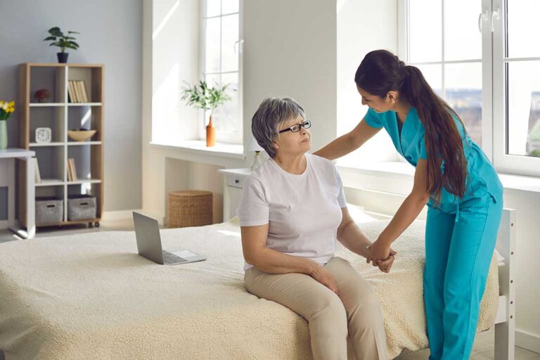 A caregiver assists an older adult with a blood pressure check in a sunlit, home-like setting.
